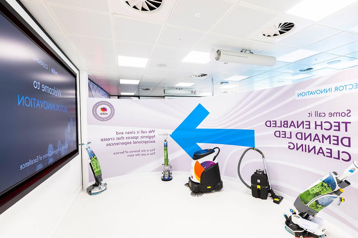 A collection of five cleaning robots and equipment at the Mitie Cleaning & Hygiene Centre of Excellence. The wall behind has text of 'Some call it tech enabled demand led cleaning'